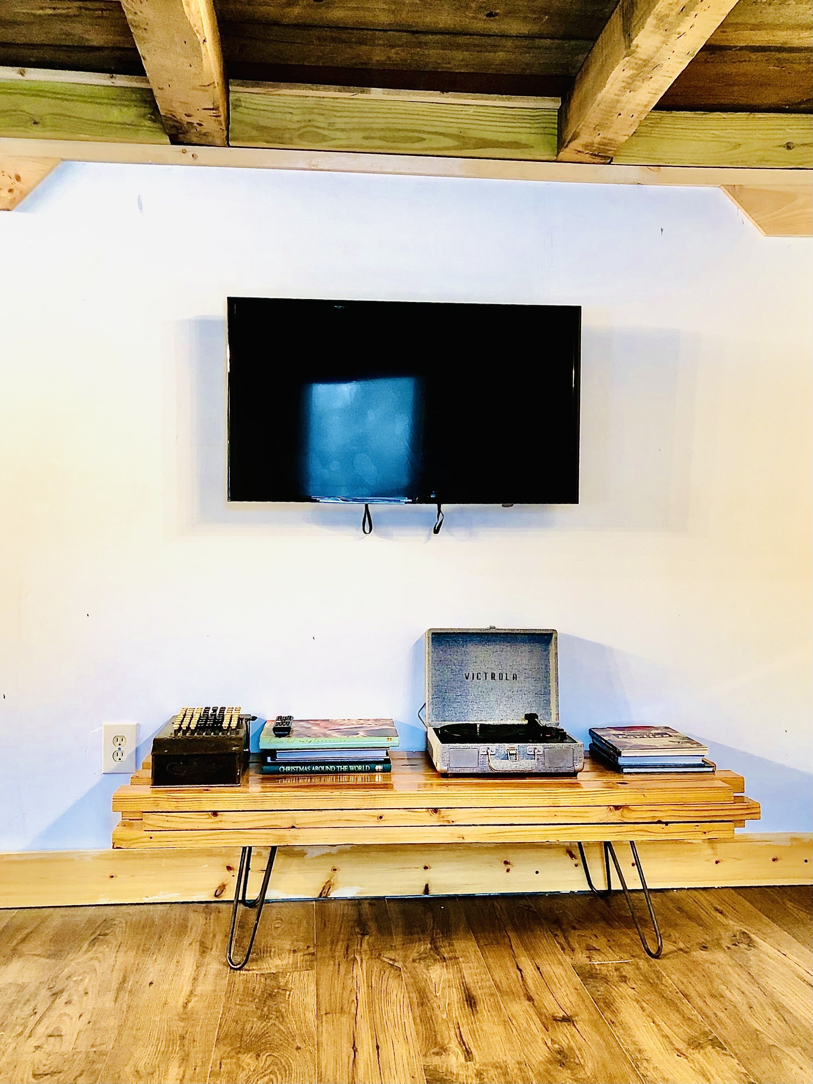 A television mounted on a white wall, a wooden table with books and electronic devices, and a metal box labeled 'VICTROLA' on the floor.