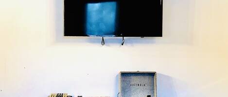 A television mounted on a white wall, a wooden table with books and electronic devices, and a metal box labeled 'VICTROLA' on the floor.