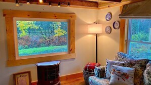 Una sala de estar acogedora con un ventanal grande, suelo de madera, un sofá con cojines estampados, una lámpara y un diploma enmarcado en la pared.