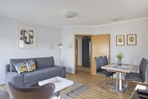 A modern living room with a grey sofa, a coffee table, and a dining area with a table and chairs. - Balcony, free use of the AHOI adventure pool and sauna - Villa Karola FeWo 19 - (Göhren)