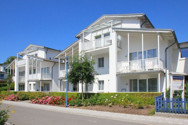 A multi-story hotel with balconies, a garden with pink flowers, and a clear blue sky.