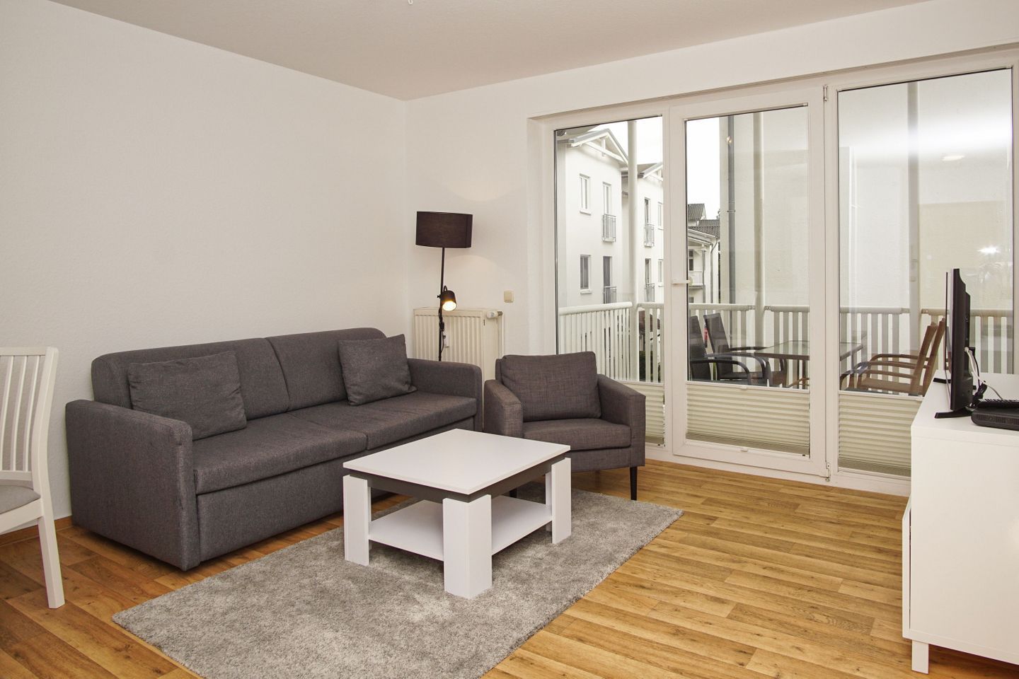 Ein modernes Wohnzimmer mit grauem Sofa, einem weißen Couchtisch und einem grauen Sessel. Ein Fernseher steht auf einem Lowboard, und es gibt einen Balkon mit Blick auf Gebäude.