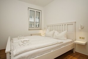 Ein weißes Bett mit Kopfteil, ein Fenster mit Blick auf einen Balkon, ein Nachttisch mit Lampe und ein kleines Regal mit Lampe.