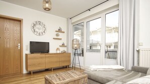 A modern living room with a wooden cabinet, a television, a wicker ottoman, and a large window with a view of buildings.