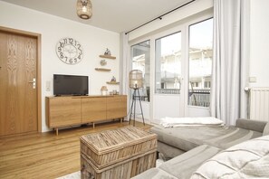 A modern living room with a wooden cabinet, a television, a wicker ottoman, and a large window with a view of buildings.