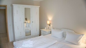 A hotel room with a large white wardrobe, a bed with white linens, a bedside table with a lamp, and a nightstand.