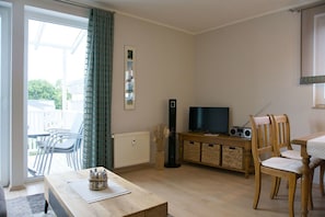 Una sala de estar con televisor, muebles de madera, una mesa de comedor y vistas a través de un ventanal.