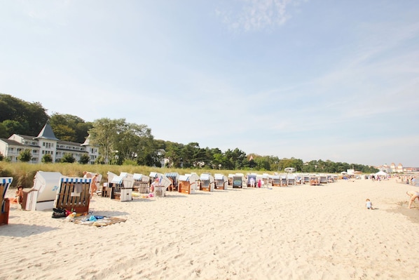 A beach with rows of beach chairs and umbrellas, a large building in the background, and a clear sky. - Directly on the beach promenade, balcony with sea view - Aparthotel Ostsee FeWo 35 (Binz)