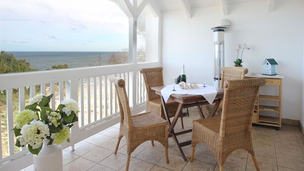 A covered patio with wicker chairs, a table with a white tablecloth, a bottle of wine, and a small house model.