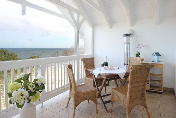 A covered patio with wicker chairs, a table with a white tablecloth, a bottle of wine, and a small house model. - Directly on the beach promenade, balcony with sea view - Aparthotel Ostsee FeWo 35 (Binz)