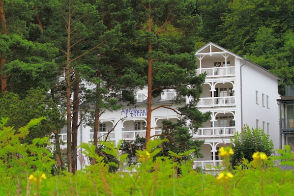 A white multi-story building with balconies, surrounded by dense green trees.