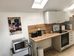 A compact kitchen with a microwave, stove, and a small sink. There's a wooden countertop, a white cabinet, and a picture of pots on the wall.