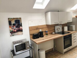 Une cuisine compacte équipée d’un four à micro-ondes, d’une cuisinière et d’un petit évier. On y trouve un plan de travail en bois, un meuble blanc et une image représentant des casseroles accrochée au mur.
