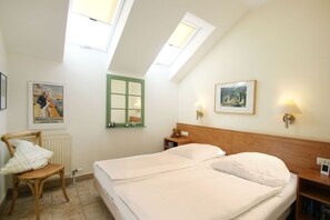 Une chambre avec deux lits, une tête de lit en bois, une table de chevet, une chaise, un radiateur, une fenêtre avec un cadre vert et deux lampes fixées au mur.