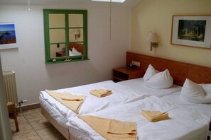 Une chambre d’hôtel avec un grand lit, deux tables de chevet, une petite table, une fenêtre encadrée de vert, une lampe fixée au mur et un tableau encadré.