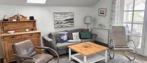 Een gezellige woonkamer met een bank, twee fauteuils, een houten bijzettafeltje, een open haard en een groot raam.