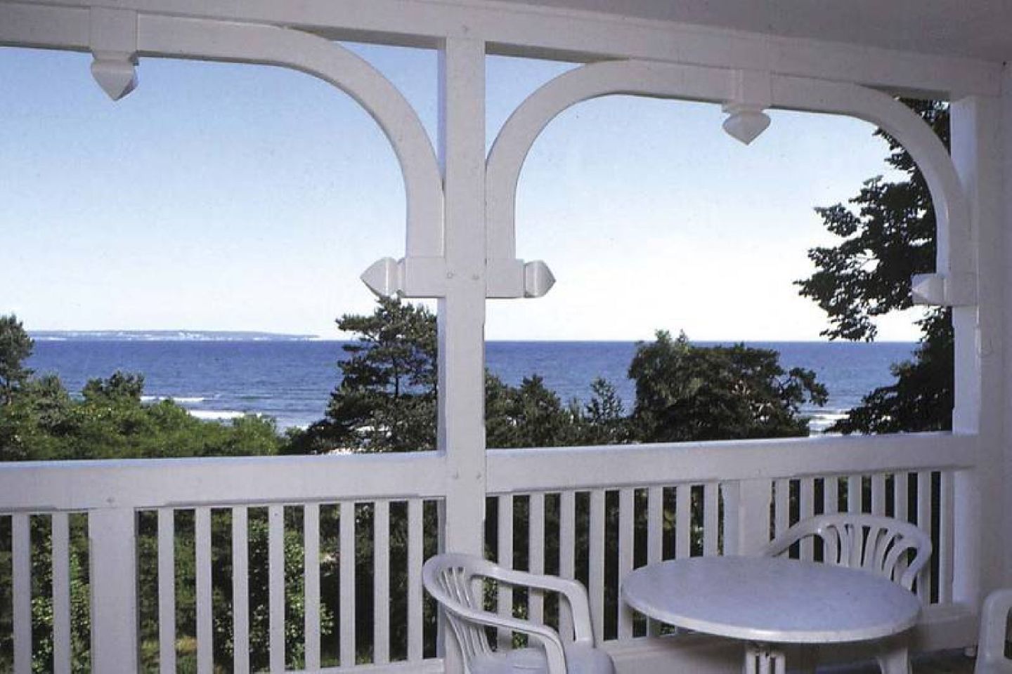Uma área externa coberta com vista para o mar, grades brancas e uma mesa redonda com cadeiras.