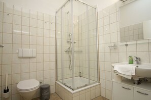 Un baño con ducha de vidrio, inodoro, cubo de basura, lavamanos con toalla y espejo.