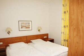 Une chambre d’hôtel avec une tête de lit en bois, deux tables de chevet, un lit avec du linge de lit blanc, deux lampes fixées au mur et un tableau encadré accroché au mur.