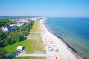 An aerial view of a beach with a row of beach umbrellas and sun loungers, a pier extending into the sea, and a row of buildings along the shore.