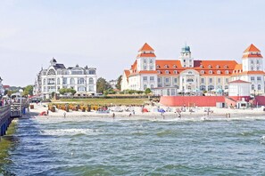 Un bâtiment situé sur la côte, doté d’une tour surmontée d’un toit rouge et d’une façade blanche, et jouxtant une plage où se trouvent des personnes et des parasols.