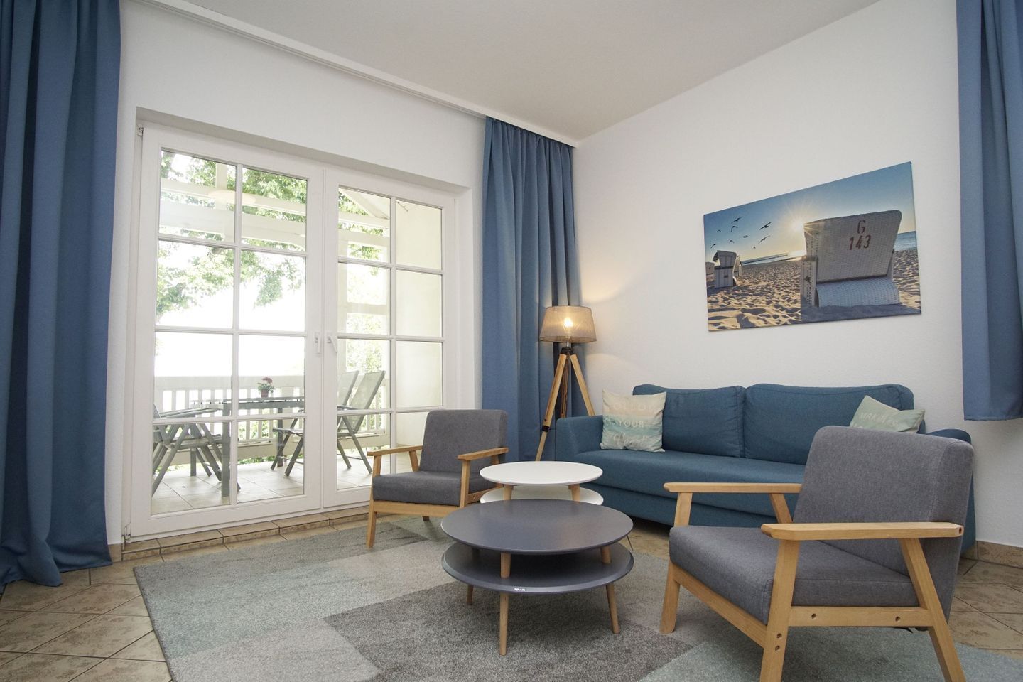 A living room with a sofa, two armchairs, a small round table, and a floor lamp. There is a large window with curtains, a painting on the wall, and a view of the outdoors.