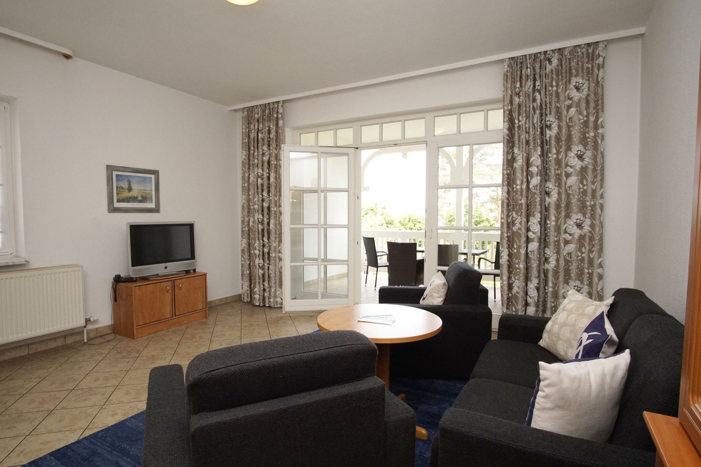A living room with a sofa, a TV on a wooden cabinet, a dining table with chairs, and a large window with curtains.