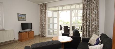 A living room with a sofa, a TV on a wooden cabinet, a dining table with chairs, and a large window with curtains.