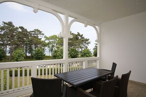 Eine überdachte Terrasse mit Tisch und Stühlen, weißen Geländern und Blick auf Bäume und ein Gewässer.