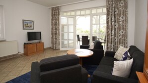 A living room with a sofa, a TV on a wooden cabinet, a dining table with chairs, and a large window with curtains.