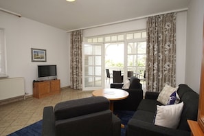A living room with a sofa, a TV on a wooden cabinet, a dining table with chairs, and a large window with curtains.