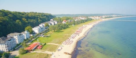 An aerial view of a coastal area with a beach, buildings, and a forested hillside.