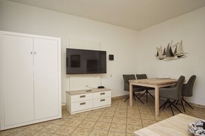 Une pièce comprenant une table à manger en bois, deux chaises, un téléviseur fixé au mur, une armoire blanche et une décoration murale représentant un navire.