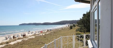A beach view from a balcony with a railing, a pier in the distance, and a grassy area with beach chairs.