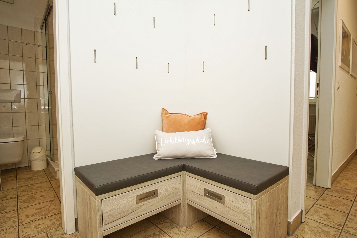 Eine Eckbank mit Staufächern und Sitzkissen mit Aufschrift, in einem Raum mit Fliesenboden und einem sichtbaren Badezimmer.