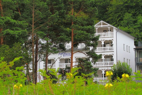Un immeuble blanc à plusieurs étages, doté de balcons, entouré d’arbres verdoyants et luxuriants.