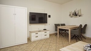 Ett rum med ett matbord i trä, två stolar, en tv som är monterad på väggen, en vit garderob och en dekorativ väggdekoration i form av ett skepp.