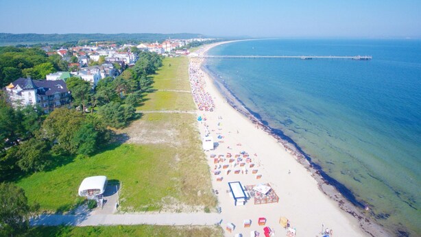 Vista aérea de uma praia com uma fileira de guarda-sóis e espreguiçadeiras, um píer que se estende para o mar e uma fileira de edifícios ao longo da costa.