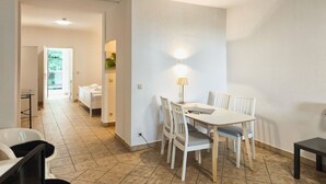 Uma sala de jantar moderna e bem iluminada, com mesa e cadeiras, um abajur e vista para uma cozinha e uma varanda.