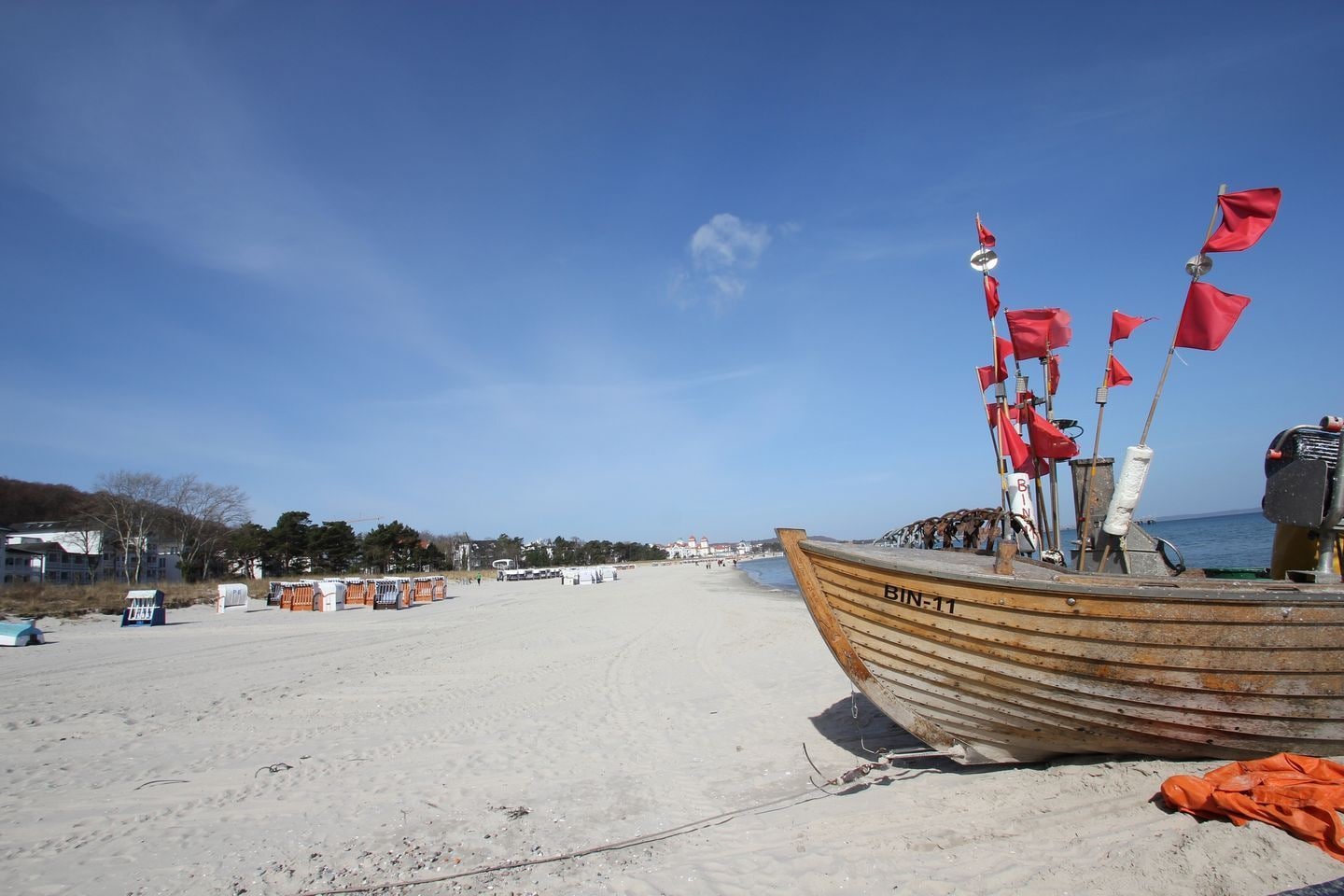 Ein Strand mit einem hölzernen Boot namens BIN-11, einem sandigen Ufer und Gebäuden im Hintergrund.