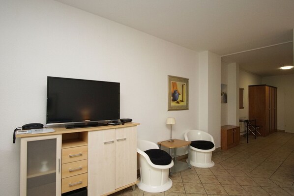 Habitación de hotel con una televisión sobre un mueble de madera, dos sillas blancas con cojines negros, una mesita con una lámpara y un cuadro en la pared.