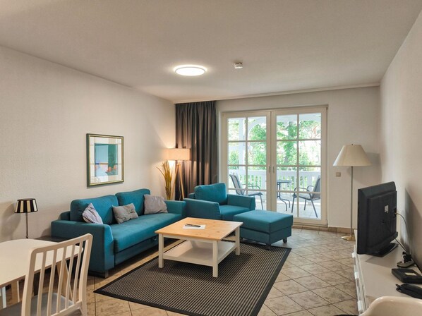 A modern living room with a blue sofa set, a wooden coffee table, and a TV. There is a large window with a view of greenery outside.