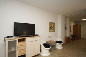 Camera d'albergo con televisione su un mobile in legno, due poltrone bianche con cuscini neri, un tavolino con una lampada e un quadro appeso al muro.