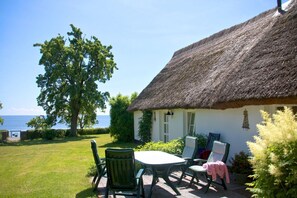 A thatched-roof cottage with a table and chairs in the garden, overlooking a body of water. - Beachfront idyll by the sea, private bathing area - Gutshof Ostseeblick FeWo 01 - (Putbus)