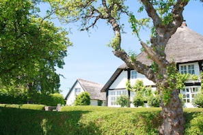 A traditional thatched-roof house with a white exterior and black trim, surrounded by well-maintained hedges and trees. - Beachfront idyll by the sea, private bathing area - Gutshof Ostseeblick FeWo 01 - (Putbus)