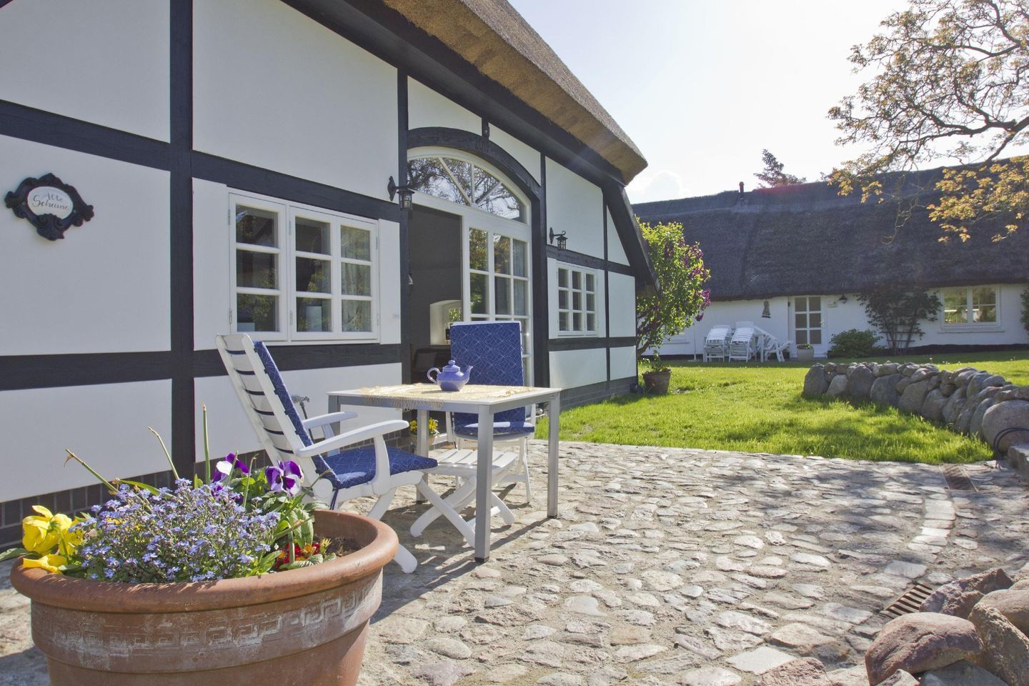 Une maison traditionnelle à colombages, dotée d’une terrasse en pierre, d’une table avec des chaises et d’un pot de fleurs.