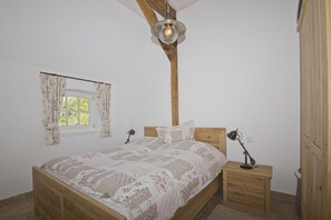 A bedroom with a wooden bed, a nightstand, a lamp, a window with floral curtains, and a wooden ceiling beam. - Idyllic seaside location close to the beach, private bathing area - Gutshof Ostseeblick FeWo 03 - (Putbus)