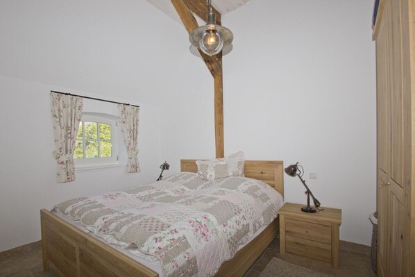 A bedroom with a wooden bed, a nightstand, a lamp, a window with floral curtains, and a wooden ceiling beam.