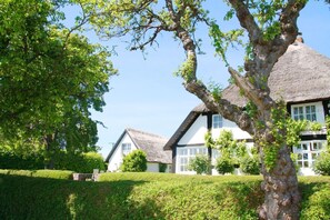 A traditional thatched-roof house with a white exterior and black trim, surrounded by well-maintained hedges and trees. - Idyllic seaside location close to the beach, private bathing area - Gutshof Ostseeblick FeWo 03 - (Putbus)