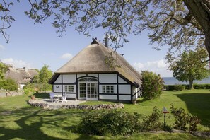 A traditional thatched-roof house with a white and black exterior, a stone pathway leading to the entrance, and a grassy garden with a small pond.